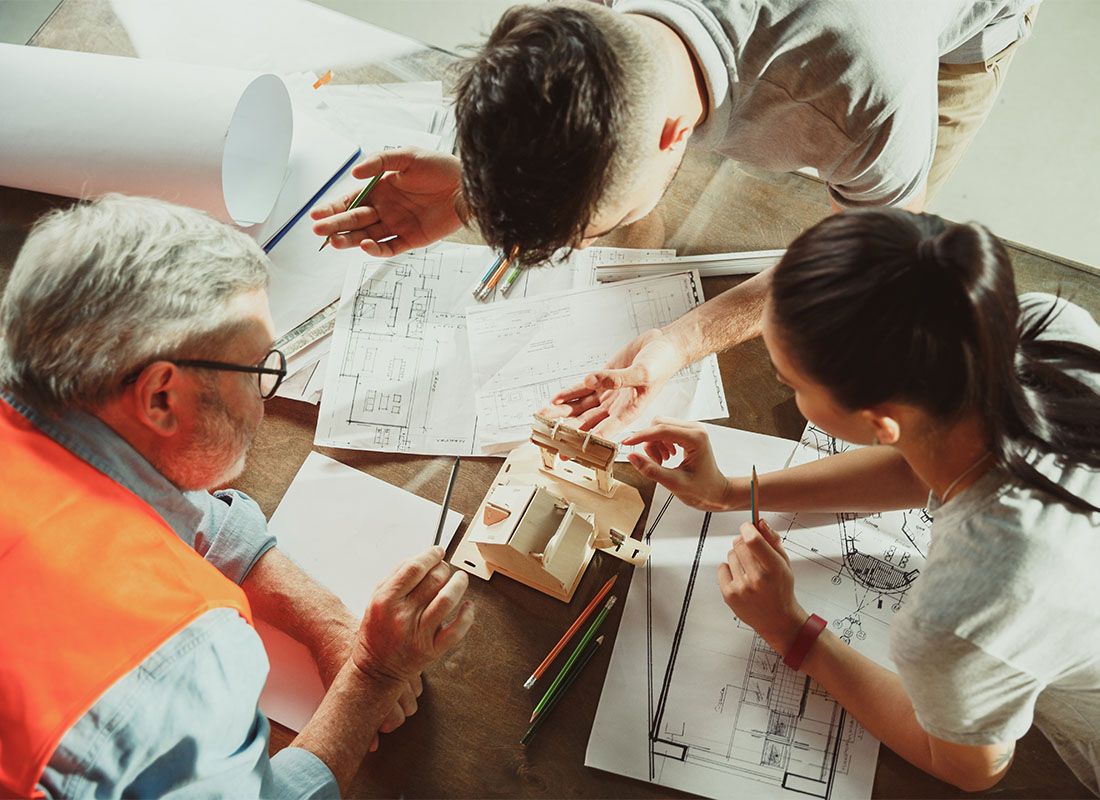 Business Insurance - View of a Contractor and Two Architects Leaning Over a Desk with New Building Blueprints and a Wooden Model During a Business Meeting