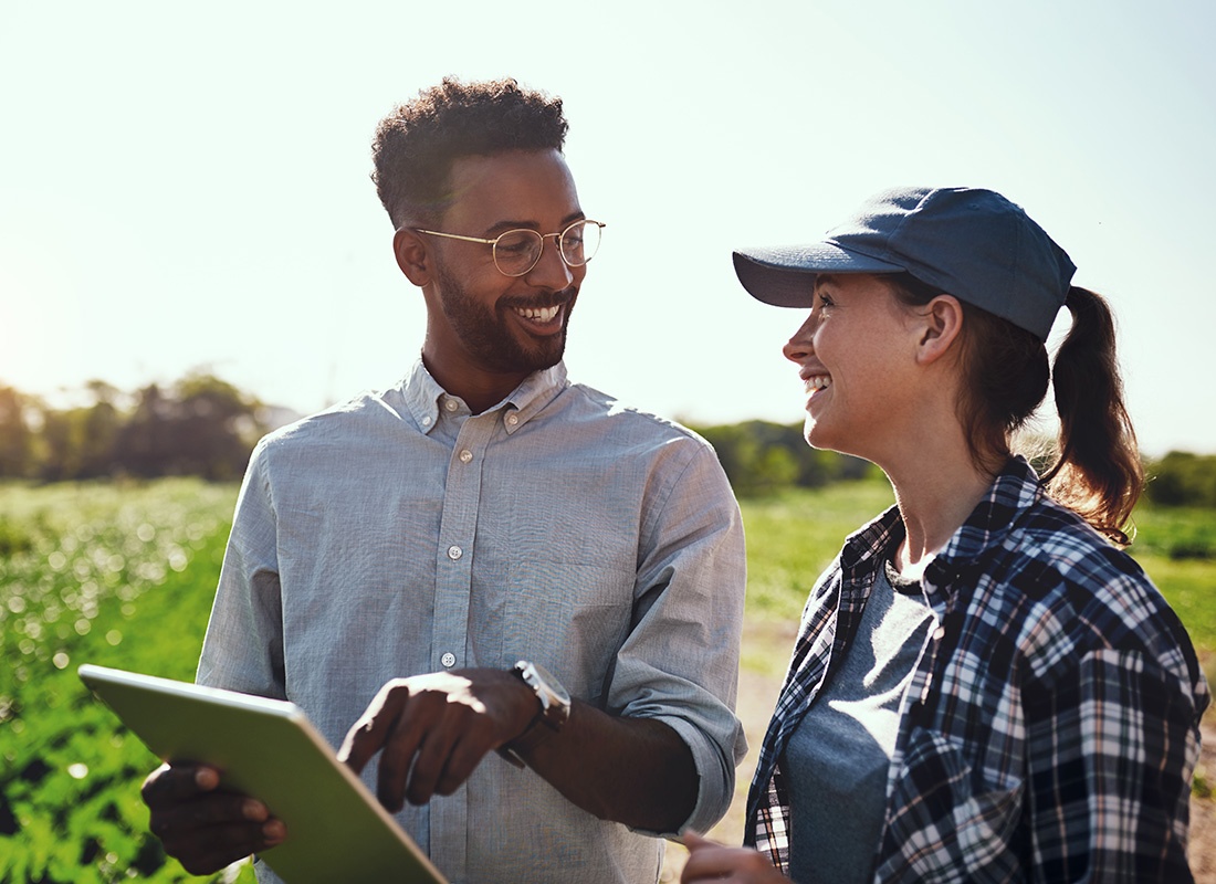 Insurance Solutions - Cheerful Young Male and Female Farmers Discussing Agriculture While Standing on a Farm Field on a Sunny Day with a Tablet