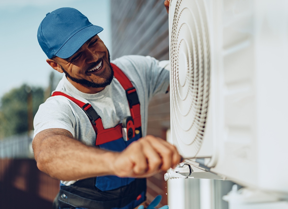 Insurance by Industry - Closeup Portrait of a Smiling HVAC Technician Working on Fixing an Outdoor Air Conditioning Unit for a Residential Home
